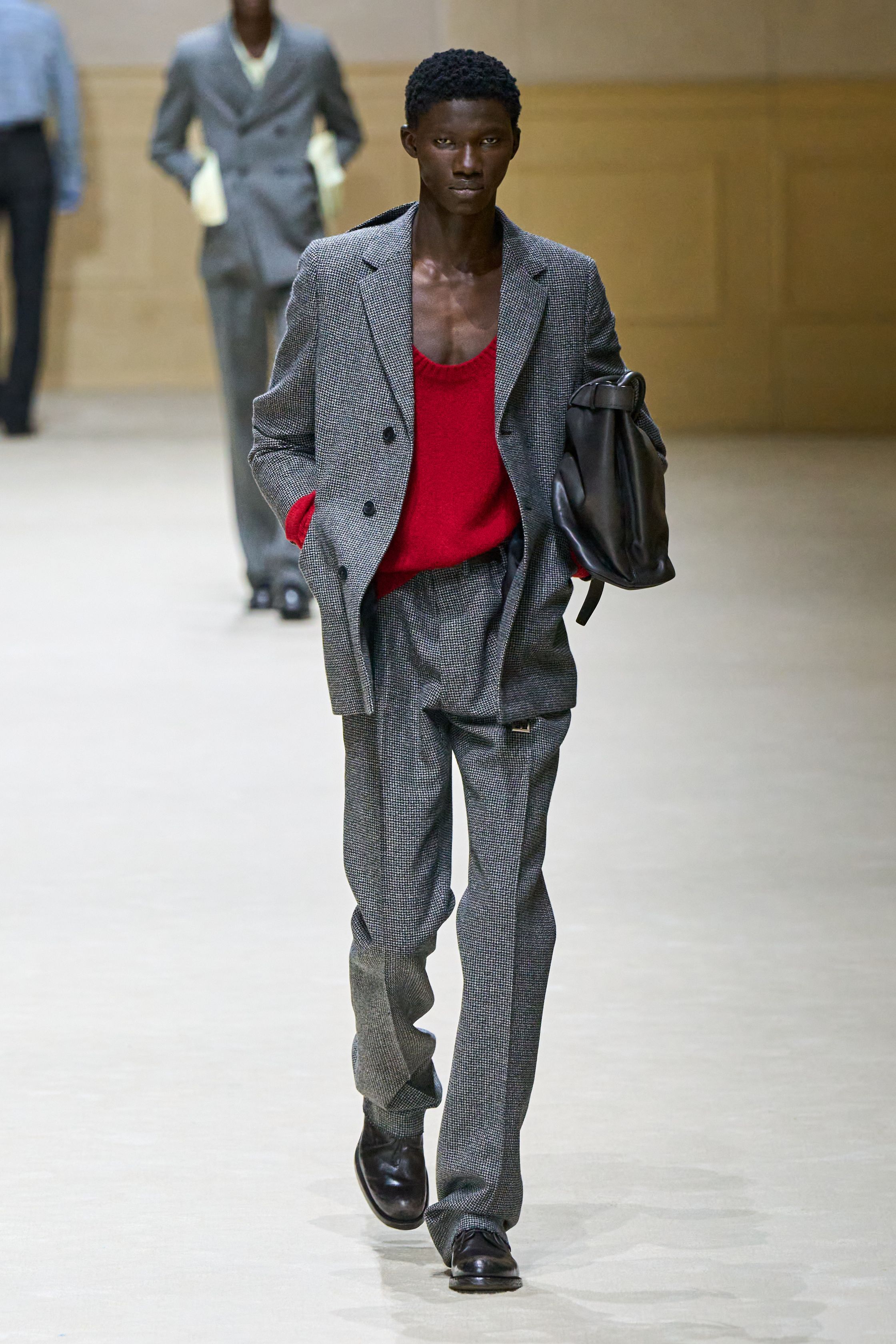 Prada 2026秋冬继续以“制服感”为切入口，重构男性权力与脆弱性的关系。修身西装、直筒大衣与功能性外套在高度理性的廓形中出现，却通过红色针织、围巾与内搭打破秩序。系列在严肃与松动之间反复摇摆，呈现一种当代男性在社会角色与个体情绪之间的张力。这是Prada一贯的冷静叙事，也是其思想性持续领先的关键简-顽石频道趋势研究